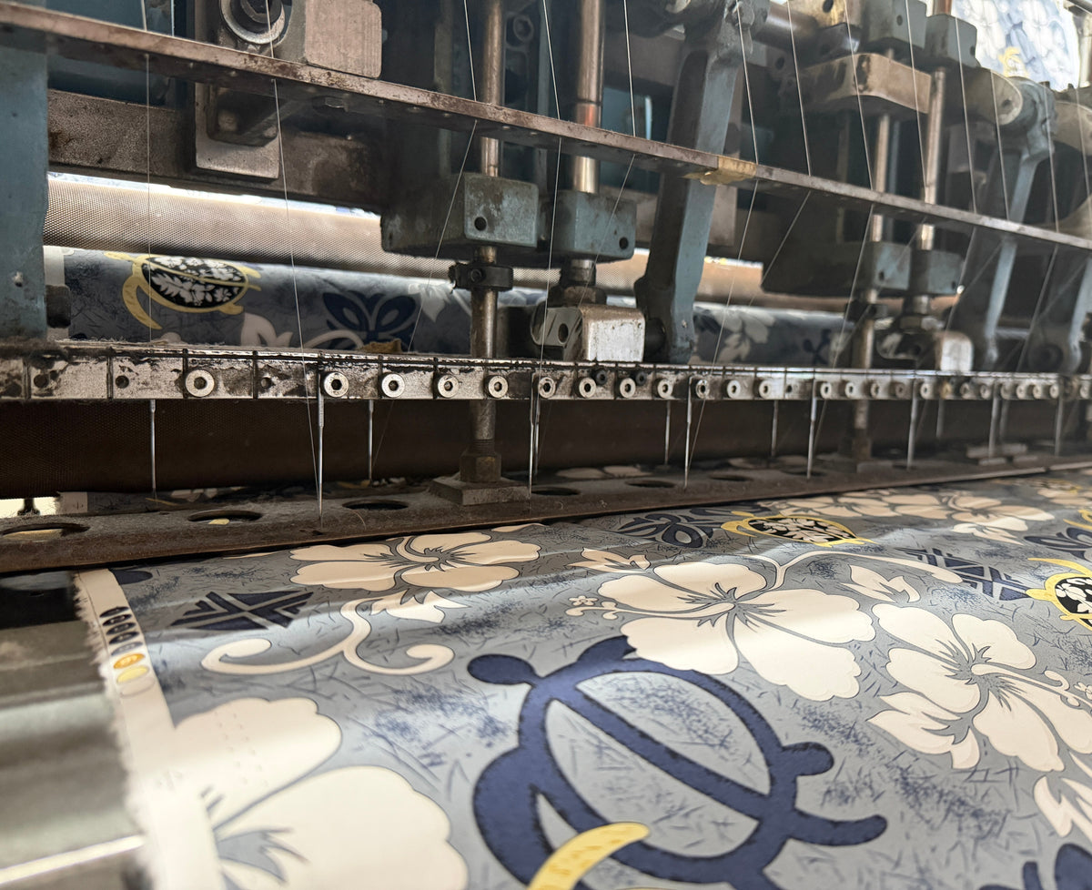 A close-up view of a patterned fabric or wallpaper being produced by a large industrial loom machine. The textile features a blue, beige, and yellow floral and scroll design.
